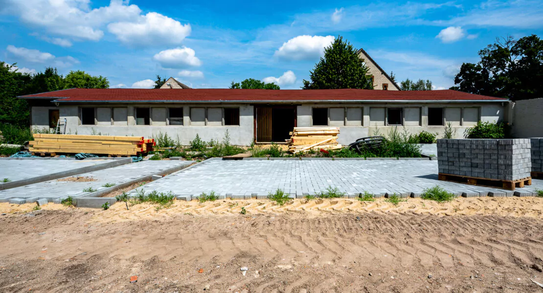 Ein graues, langgestrecktes, einstöckiges Haus mit einem roten, schrägen Flachdach und vielen Fenstern, das gerade renoviert wird.