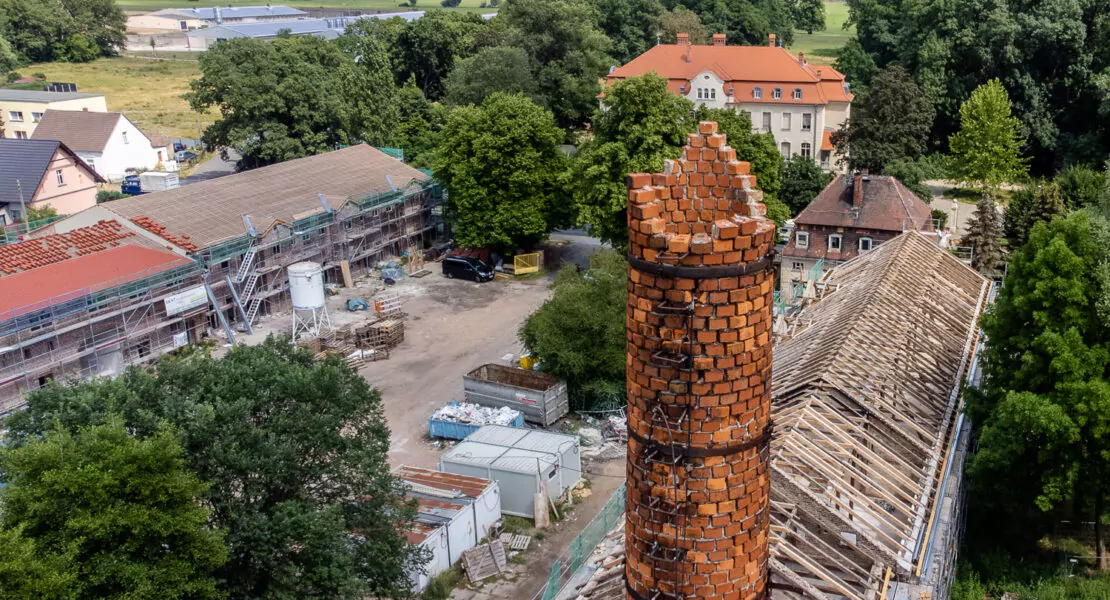 Ein Schornstein, der oben zerfällt, im Hintergrund eine Villa und viele Felder.