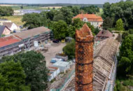 Ein Schornstein, der oben zerfällt, im Hintergrund eine Villa und viele Felder.