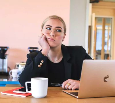 Frau in Blazer und frustriertem Blick sitzt an einem Tisch hinter einem Laptop