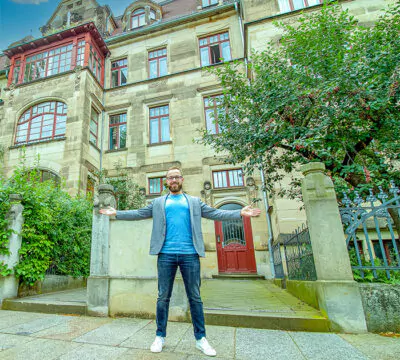 Fabian Schuster steht mit ausgebreiteten Armen vor einem Mehrfamilienhaus