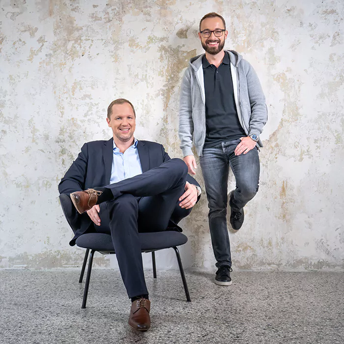 Marcus Jurowski (links) sitzt auf einem Stuhl, Fabian Schuster (rechts) lehnt hinter ihm an einer Wand