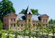 Kleines Schloss hinter einer Hecke mit Turm und drei Stockwerken.