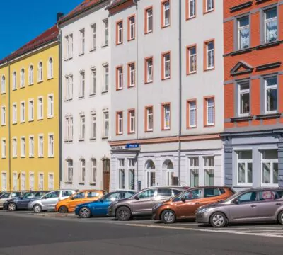 Eine Straße mit älteren Immobilien. Vor den Immobilien stehen geparkte Autos.