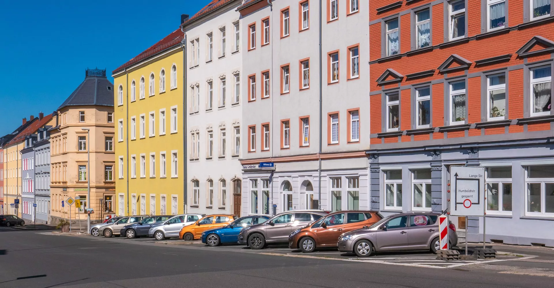 Eine Straße mit älteren Immobilien. Vor den Immobilien stehen geparkte Autos.